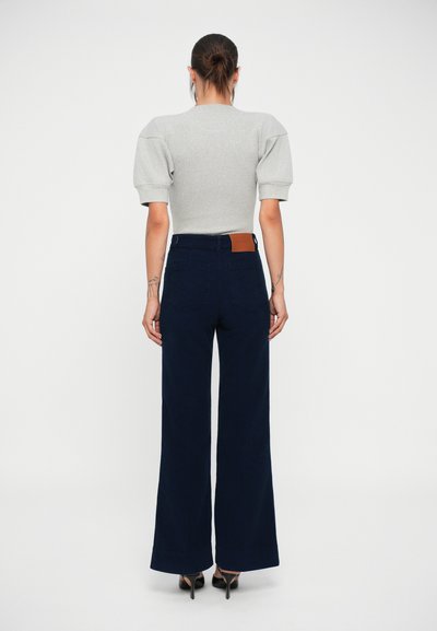 Top gri cu mâneci bufante, asortat cu pantaloni largi cu talie înaltă, de culoare bleumarin, care au buzunare pe spate și un accent din piele maro. Material tricotat texturat.
