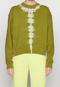 Oliwkowa bluza z wzorem tie-dye w białym kolorze, okrągły dekolt, opadające ramiona i ściągacz u dołu, w zestawie z jasnoniebieskimi spodniami.