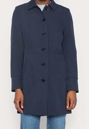 Manteau bleu marine boutonné avec col, manches longues et poches latérales porté sur un jean bleu clair.