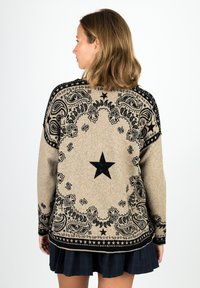 Woman with shoulder-length hair wears beige sweater with black star and paisley pattern, paired with dark pleated skirt, facing away.