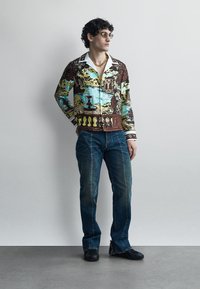 Hombre con cabello rizado que lleva una camisa de manga larga con estampado, vaqueros azules, zapatos negros y gafas de sol, de pie frente a una pared lisa.