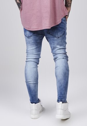 Personne debout les mains dans les poches, portant un jean bleu délavé, une chemise rose, des baskets blanches et des tatouages visibles sur les bras.