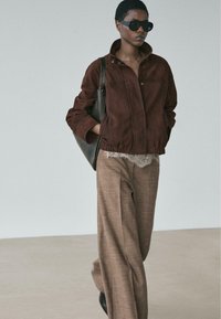 Braune Wildlederjacke mit Kragen, kombiniert mit braunen Weithose. Das Modell trägt eine schwarze Tasche und große dunkle Sonnenbrillen.