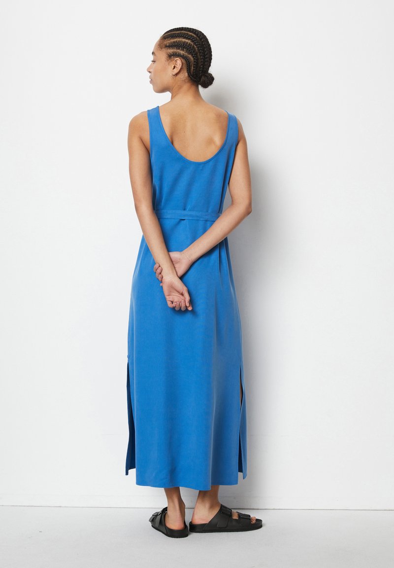 Blaues ärmelloses Baumwollkleid mit tiefer Rückenpartie, seitlichen Schlitzen und einer Taillenbindung. Model trägt schwarze Sandalen und steht mit dem Rücken zur Kamera.