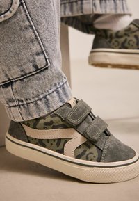 Zielone zamszowe sneakersy z beżowymi akcentami, posiadające wzór w kamuflażu, rzepy oraz teksturowaną podeszwę, w połączeniu z dżinsami.