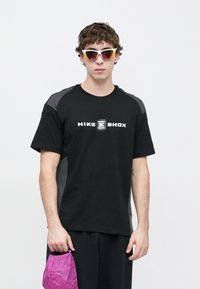 Camiseta preta com o logotipo "NIKE SHOX" em branco, detalhes cinzentos nas mangas, combinada com calções escuros e segurando uma pequena bolsa rosa.