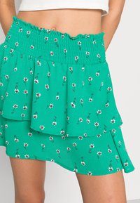 Satiro verde com padrão floral, apresentando uma cintura franzida, camadas de frufrus e pequenos padrões de flores brancas e vermelhas em um tecido leve.