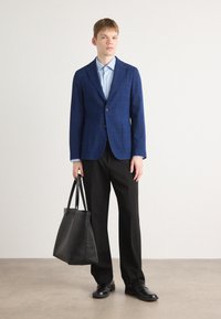 Modra karo opravljena blazer preko svetlo modre srajce, v kombinaciji s črnimi hlačami in čevlji. Črna torba se drži v eni roki, preprosta ozadje.