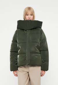Mørkegrønt puffjakke med høj krave, skinnende materiale, quiltet mønster og en boxy pasform. Modellen har beige bukser på.