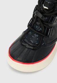 Zapatilla alta negra con parte superior de goma texturizada, lengüeta de tela estampada, acento rojo en la suela y cordones negros con cierre tipo toggle.