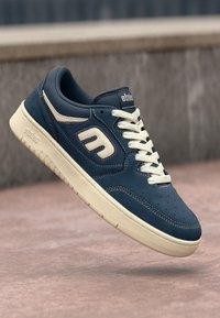 Blue suede sneaker with white accents, rounded toe, and flat lace-up design. Textured paneling and logo on the side. Cream rubber sole.