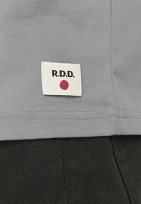 Camiseta de algodón gris con dobladillo cosido, que presenta una etiqueta blanca con "R.D.D." en negro y un punto rojo, exhibida sobre una tela oscura.