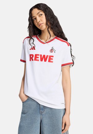 Kobieta z długimi kręconymi włosami, ubrana w białą koszulkę piłkarską z czerwonym logo REWE i emblematem drużyny, w połączeniu z niebieskimi dżinsami, stojąca z jedną ręką w kieszeni.