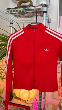 Rote sportliche Jacke mit Reißverschluss und gerippter Textur, weiße drei Streifen-Akzente an den Ärmeln und kleinem weißen Logo auf der Brust.