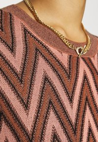 Gruba sweter w odcieniach różu i brązu z wzorem w zygzaki oraz akcentami w czarnym kolorze. Złoty łańcuszek dodaje szczegółu.