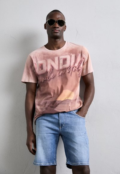 Camiseta de manga corta, color rosa claro, con "Londres Hecho en Italia" impreso en un tono más oscuro, combinada con pantalones cortos de mezclilla azul.