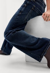 Person wearing dark blue jeans and brown shoes, sitting on a white block, with a hand resting on the block showing gold rings.