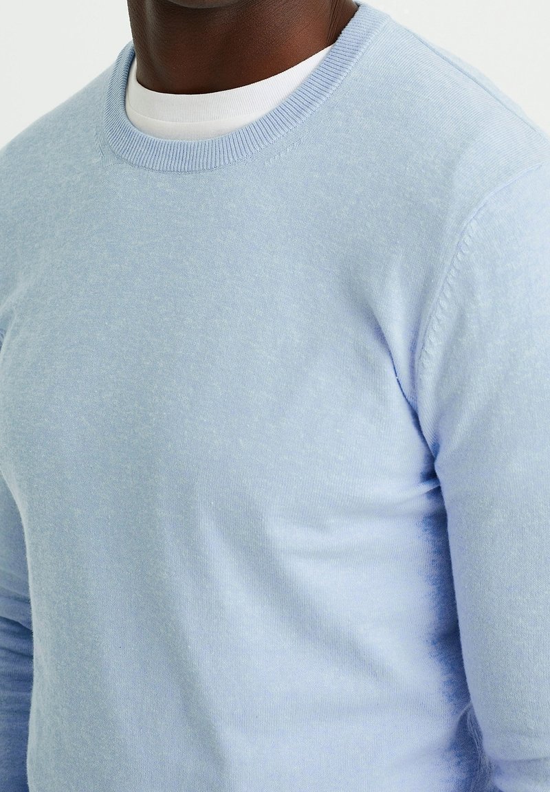 Homme portant un pull col rond bleu clair par-dessus une chemise col rond blanche, torse et cou visibles, fond neutre.