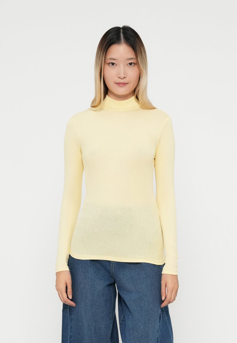 Suéter de cuello alto de manga larga en un suave color amarillo claro, confeccionado en una tela suave que presenta una forma ajustada y una textura lisa. Combinado con unos vaqueros de pierna ancha en azul.