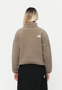 Brun fleecejakke med høy krage, prydet med en liten hvit logo på øvre bak, kombinert med ett svart cargo-skjørt.