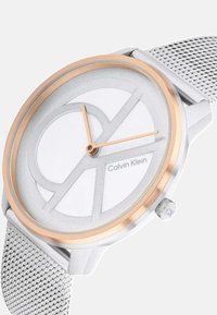 Sidabrinis ir rožių aukso tinklinis dirželis su apvaliu baltu ciferblatu, kuriame yra geometriniai raštai ir oranžinės rodyklės. Rodyklėje matomas Calvin Klein logotipas.