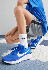 Blaue Sportschuhe mit einem strukturierten Obermaterial, schwarzen Akzenten und einem weißen Swoosh-Logo. Getragen mit weißen Socken, die ein schwarzes Logo haben.