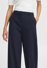 Navy široke hlače s glatkom teksturom, s detaljima nabora sprijeda, pojasom s remenom i bočnim džepovima.