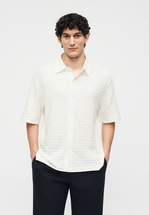 Άνδρας με σγουρά μαλλιά που φοράει λευκό υφασμάτινο κοντομάνικο πουκάμισο και μαύρο παντελόνι με τα χέρια στις τσέπες.