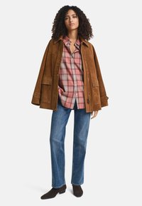 Vrouw met krullend haar, gekleed in een bruine suède jas over een roze geruit shirt, blauwe jeans en donkerbruine enkellaarzen, staande op een witte achtergrond.