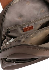 Intérieur d'un sac en tissu marron présentant une étiquette en cuir beige, plusieurs compartiments zippés et une texture lisse avec une doublure à motif subtil.