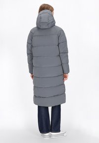Palton lung, căptușit, de culoare gri, cu glugă, având secțiuni orizontale pentru izolație. Purtați peste blugi negri din denim și adidași albi.