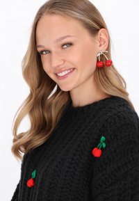 Červené náušnice vo forme čerešní so striebornými akcentmi na modele, ktorý má na sebe čierny pletený sveter s malou červenou výšivkou čerešní.