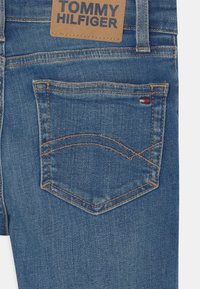 Vaqueros de mezclilla azul con una etiqueta de cuero marrón en la cintura y un bolsillo trasero cosido. Presentan un pespunte ligero y un pequeño acento rojo y blanco.