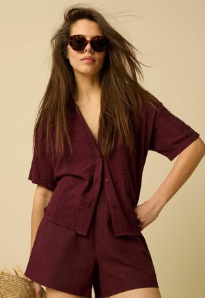 Femme aux longs cheveux portant des lunettes de soleil écaille de tortue et une chemise bordeaux texturée à boutons avec un short assorti sur un fond beige.