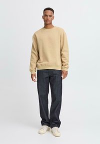 Beige crew neck sweatshirt with relaxed fit, paired with dark blue denim jeans. Model wearing off-white sneakers, standing against a plain background.