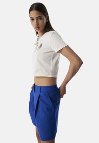 T-shirt blanc en coton coupé, avec des patches décoratifs, associé à un short en lin bleu structuré avec des plis et des poches latérales.