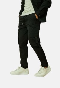 Pantalons cargo noirs avec poches latérales, fabriqués en tissu léger. Associés à des baskets blanches, mettant en avant un design ajusté.