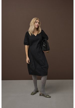 Blondínka v čiernych šatách po kolená, sivých pančuchách, nízkych hadích podpätkoch, drží veľkú čiernu kabelku na hnedom pozadí.