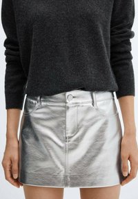 Mini jupe argentée métallique avec une coupe ajustée, poches avant et fermeture à boutons. Texture lisse et finition brillante. Portée avec un pull noir.