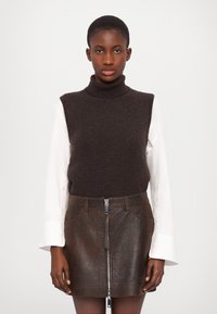 Maglione senza maniche marrone a collo alto indossato sopra una camicia bianca a maniche lunghe, abbinato a una minigonna in pelle marrone con zip frontale.