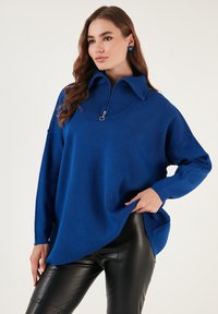 Pulover tricotat albastru cu guler înalt și închidere cu fermoar, având mâneci lungi și o croială supradimensionată, asortat cu pantaloni din piele negri.