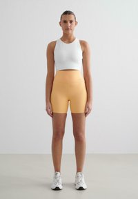 Fehér crop top és világos narancssárga, magas derekú rövidnadrág. Az anyag simának és rugalmasnak tűnik. A modellen fehér sportcipő van. Egyszerű háttér.