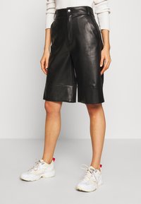 Svart läderbermuda med knappstängning fram, hög midja och sidfickor. Kombinerad med vita sportiga sneakers.