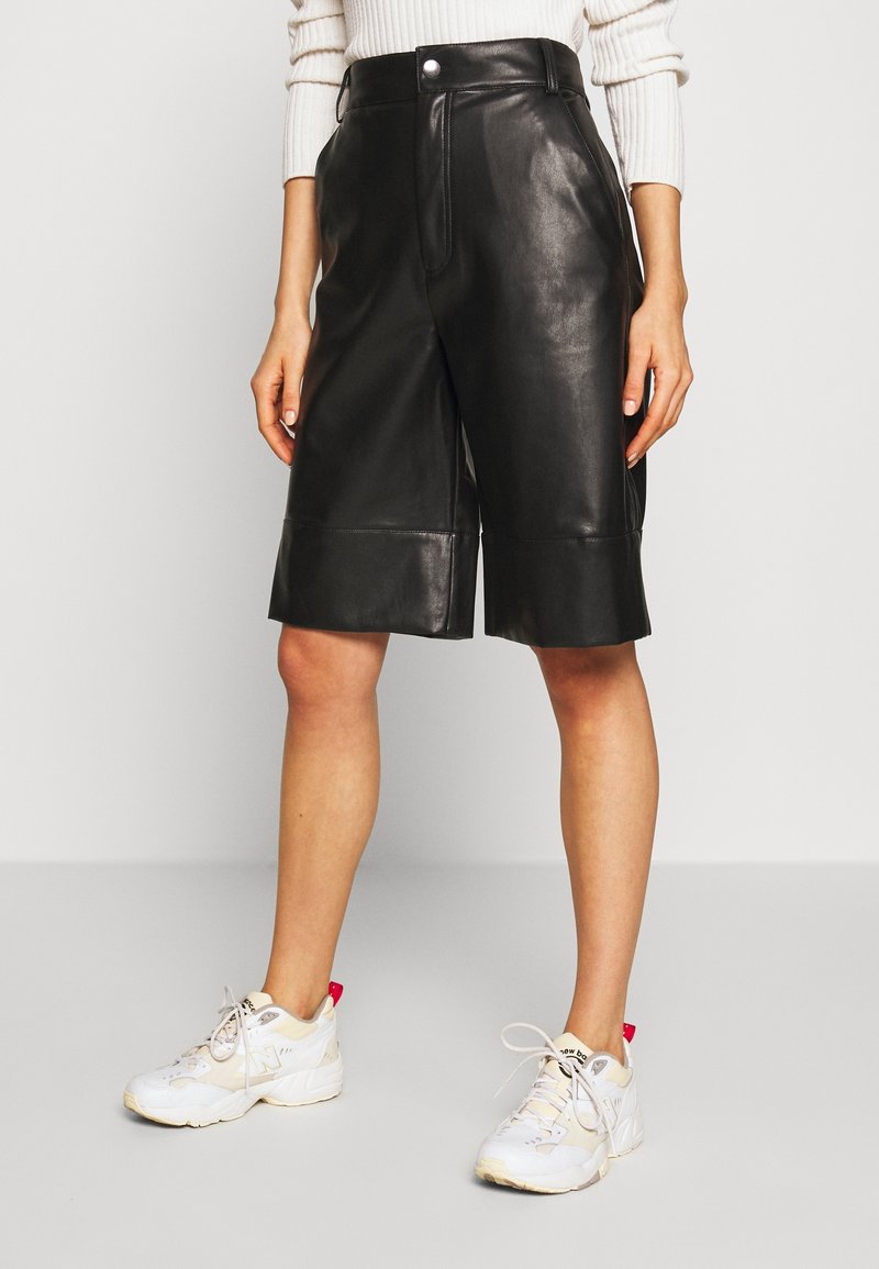 Svart läderbermuda med knappstängning fram, hög midja och sidfickor. Kombinerad med vita sportiga sneakers.