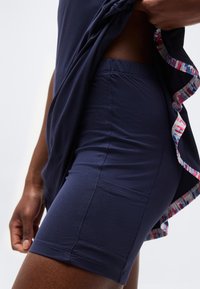 Κοντινό πλάνο ατόμου που σηκώνει μπλε σκούρο μπλουζάκι για να δείξει τα ταιριαστά ποδηλατικά σορτς σε μπλε σκούρο χρώμα με πλαϊνή τσέπη και πολύχρωμη λεπτομέρεια στο κάτω μέρος του μπλουζακιού.