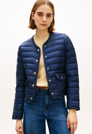 Kvinne iført en marineblå quiltet jakke med gullknapper, beige kabelstrikket genser og blå jeans mot en ensfarget hvit bakgrunn.