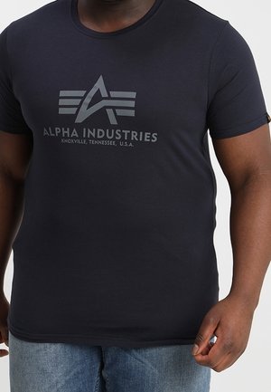 Homme portant un t-shirt noir ajusté Alpha Industries avec logo et texte, associé à un jean bleu, debout devant un fond uni.