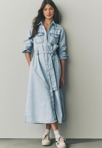 Ljusblå denimskjolklänning med långa ärmar, två bröstfickor och ett knytbälte i midjan. Har en knappstängning fram och sidoklyftor.
