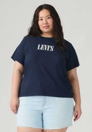 Kvinde med langt mørkt hår iført en navy Levi's t-shirt og lyseblå denimshorts, stående mod en ensfarvet lys baggrund.