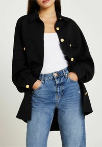 Svart vadderad skjortjacka med guldknappar, med krage, långa ärmar och en avslappnad passform, bärs över en vit linne och blå jeans.